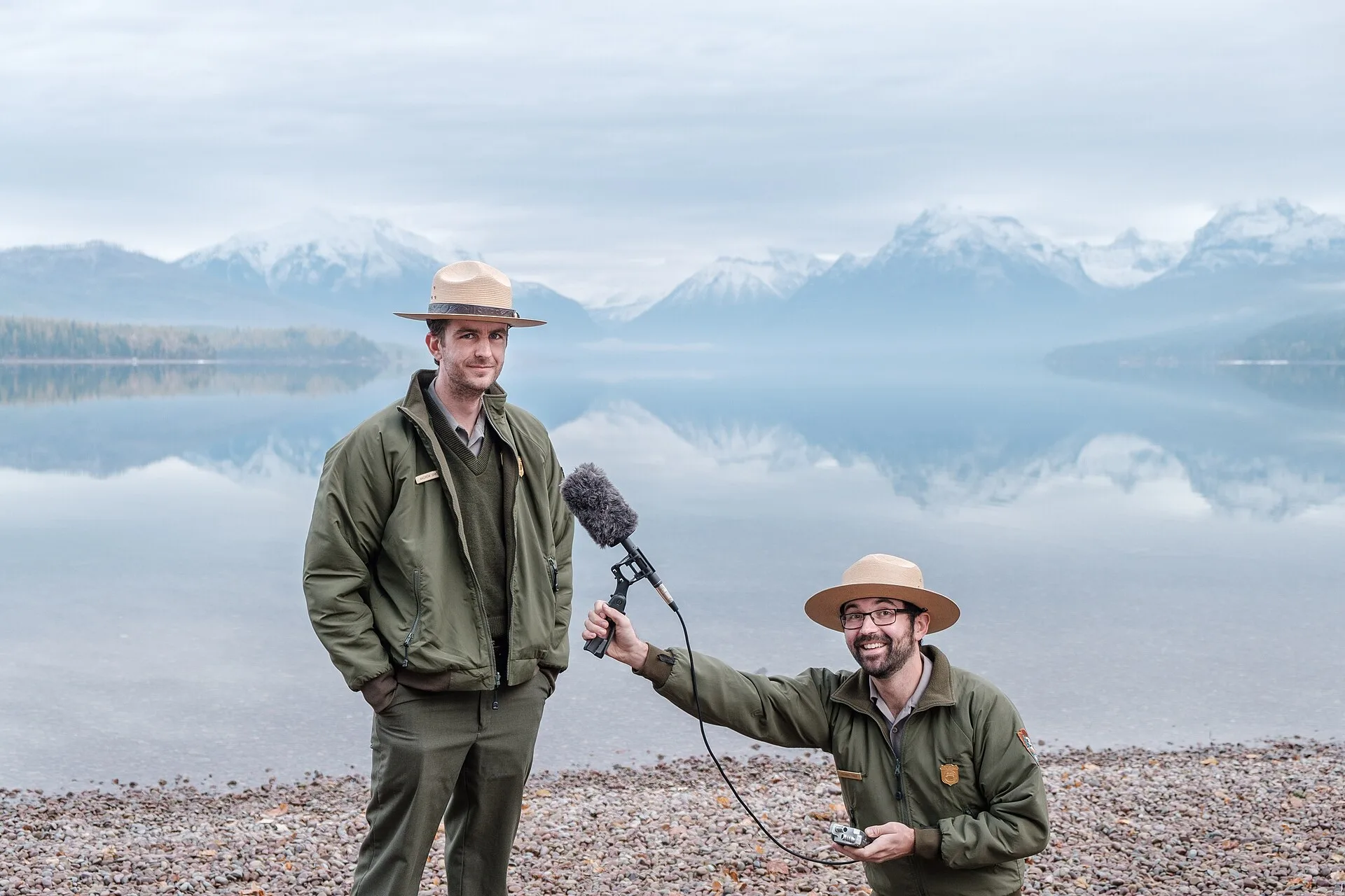 Headwaters Podcast Hosts Andrew Smith and Michael Faist posing with a shotgun mic in front of Lake McDonald.