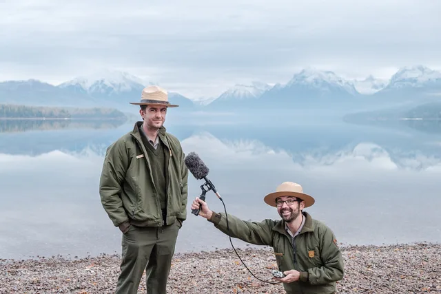 Headwaters Podcast Hosts Andrew Smith and Michael Faist posing with a shotgun mic in front of Lake McDonald. Podcast SEO buyers: who pays and what proof they want