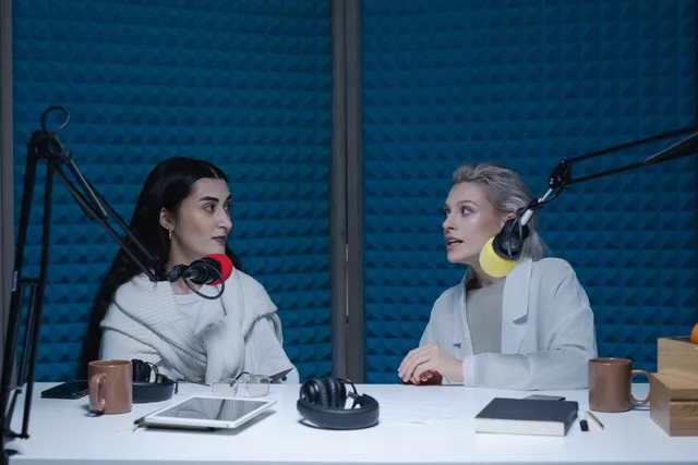 Two women engaged in conversation, recording podcast in studio with microphones and headphones. How to build a podcast SEO skills forecast for England