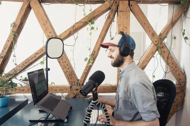 A man with headphones podcasts at a desk with a laptop and microphone stand indoors. Before you draft a podcast SEO strategy framework, score it first